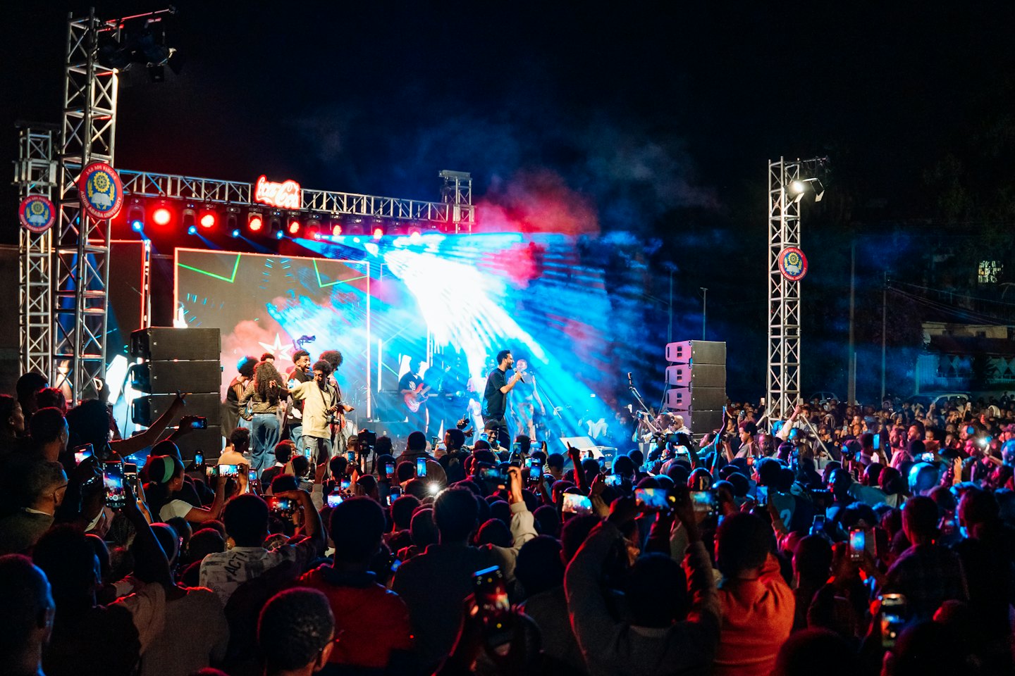 Musicians perform on stage while a crowd records with smartphones.