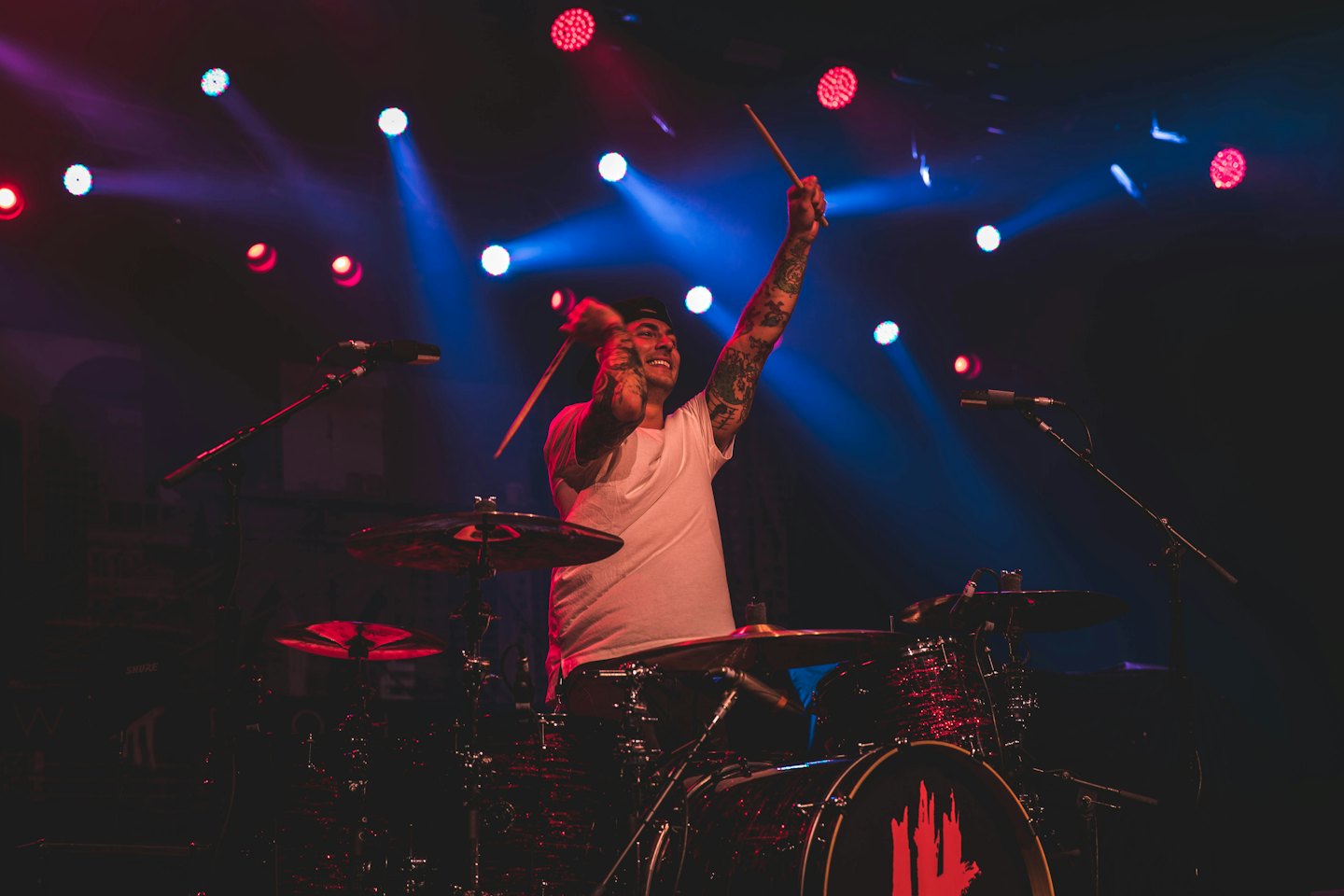 Smiling male drummer with arm tattoos raises drumsticks on a dark stage.