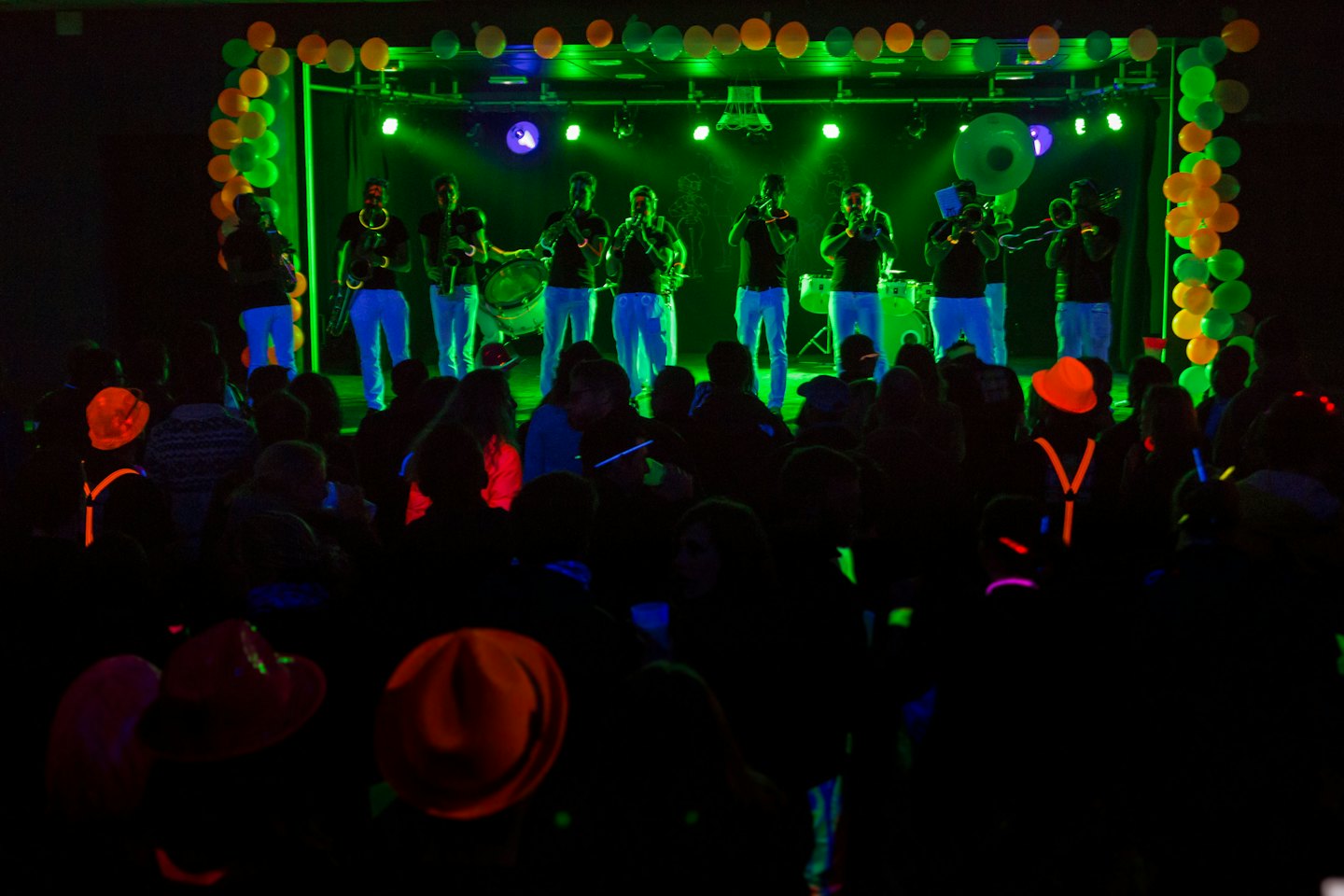 Musicians play horns and drums on a neon stage with audience.